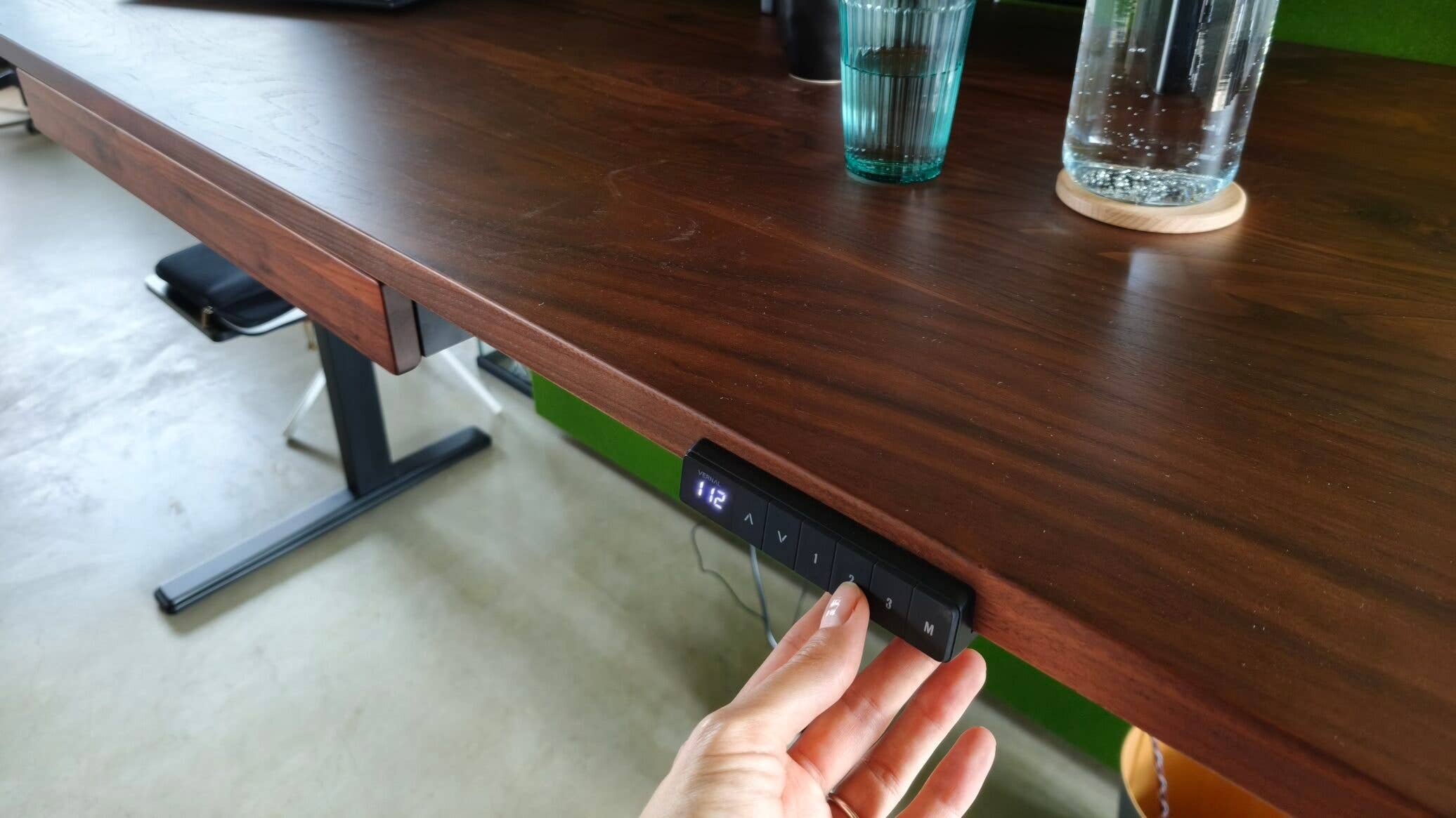 Schreibtisch, Bedienfeld zum Hoch- und Runterfahren, Wasserflasche aus Glas und Glas gefüllt mit Wasser, Laptop, Bildschirm, Hand drückt aufs Bedienfeld