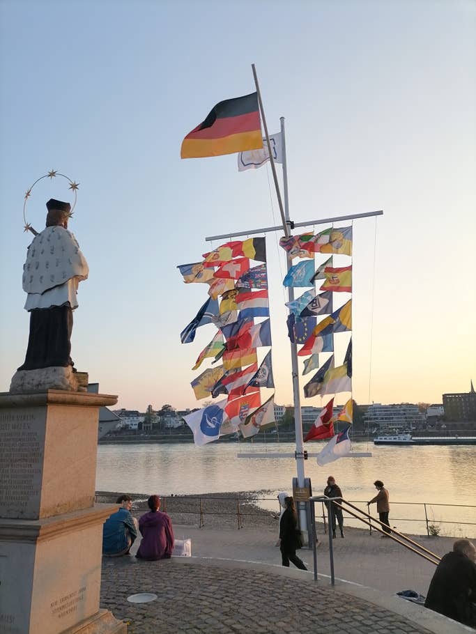 Eine Heiligenstatue und Fahnen im Vordergrund; dahinter der Rhein.