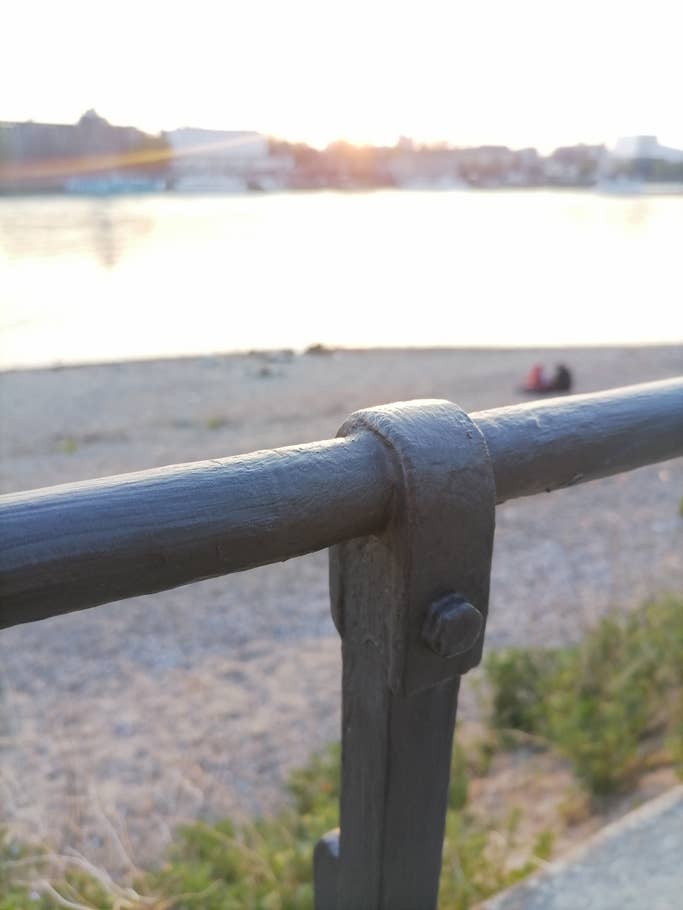 Ein Metallgeländer im Fokus mit Wasser und Strand im Hintergrund.