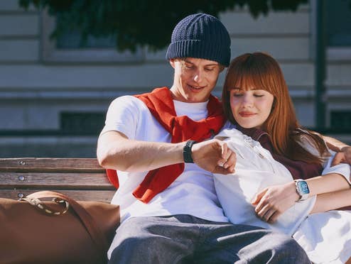 Ein Mann und eine Frau sitzen auf einer Bank und blicken auf die Smartwatches auf ihren Handgelenken.