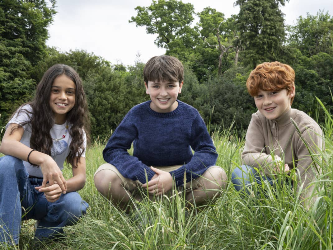 Drei Kinder lächeln, während sie im Gras sitzen, mit Bäumen im Hintergrund.