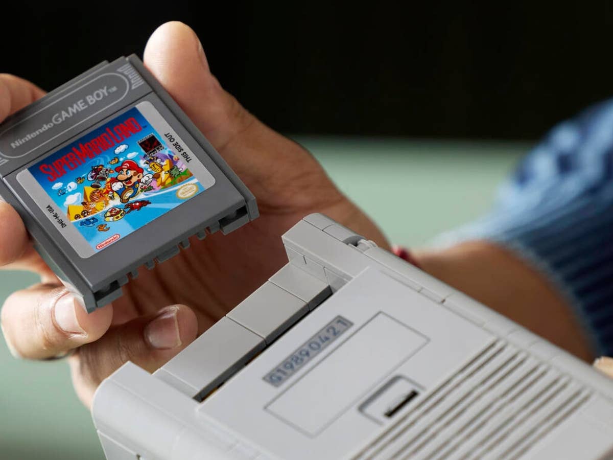 Eine Person hält eine Game Boy-Cartridge mit der Bezeichnung 'Super Mario' über einer Game Boy-Konsole mit der Cartridge 'Zelda'.