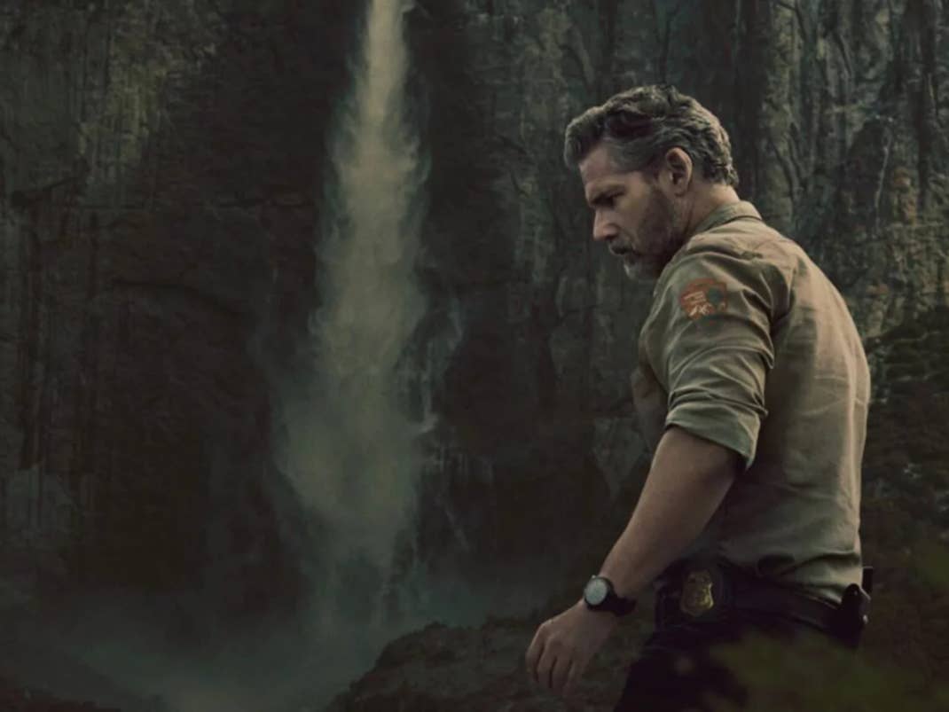 Ein Mann steht in der Nähe eines Wasserfalls, trägt ein beiges Hemd, mit einer felsigen Klippe im Hintergrund.