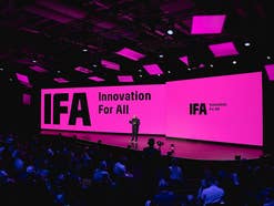 Ein Redner steht auf einer großen Bühne vor zwei pinkfarbenen Leinwänden mit der Aufschrift „IFA Innovation For All