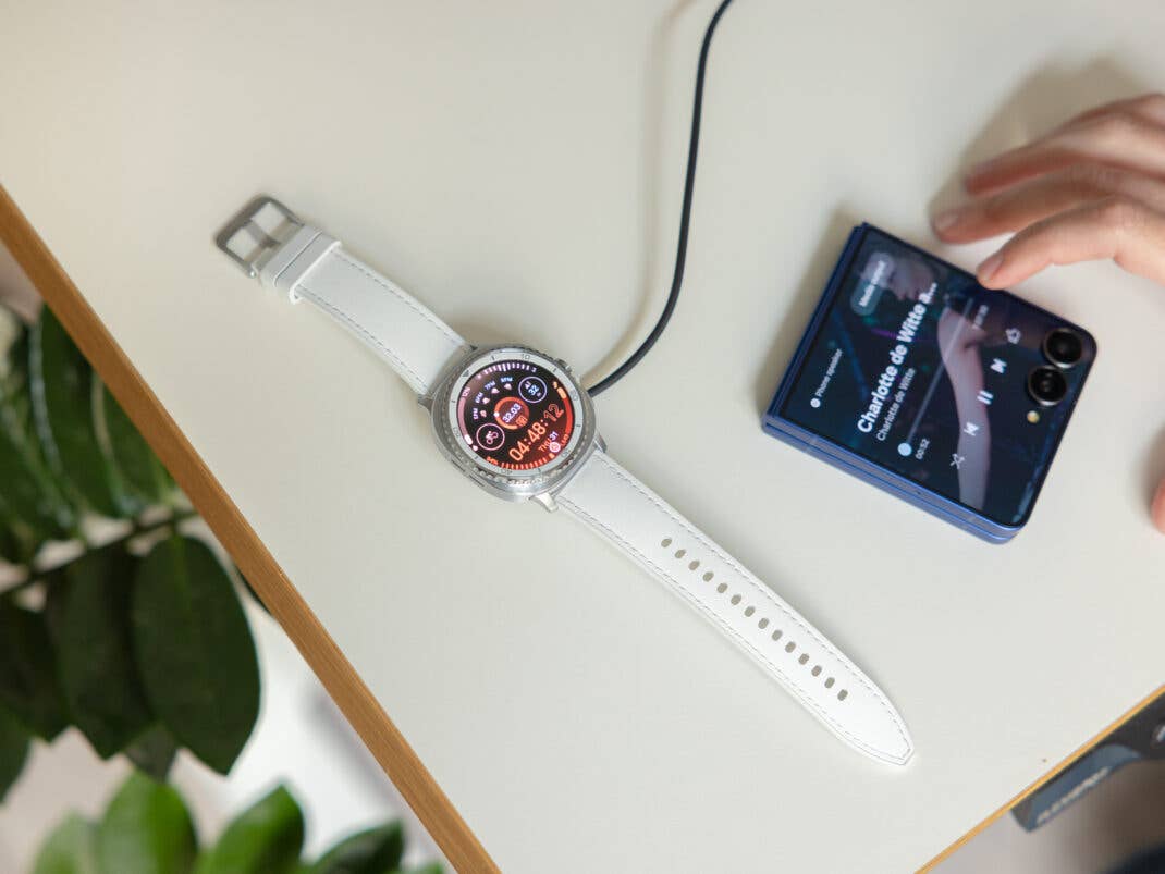 Samsung Galaxy Watch 8 Classic auf einem Tisch, lädt, neben einem Smartphone, das Musik abspielt.