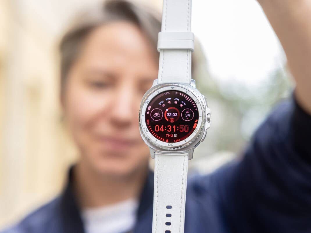 Eine Person hält eine Samsung Galaxy Watch 8 Classic, die ein rotes Interface mit Uhrzeit und Fitnessstatistiken zeigt.
