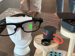 Eine schwarze Smart-Brille von INMO Technology steht auf einem weißen Kopfmodell, daneben liegen ein kleiner schwarzer Lautsprecher, ein Ring und ein aufgeklapptes Ladecase auf Holzständern.