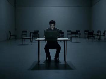 Eine Person sitzt allein an einem Tisch mit einem Laptop in einem dunklen, leeren Raum.