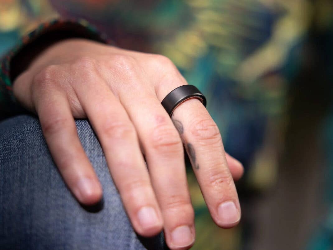 Eine Nahaufnahme einer Hand, die einen schwarzen Oura-Ring am Finger trägt und auf einem Knie ruht.