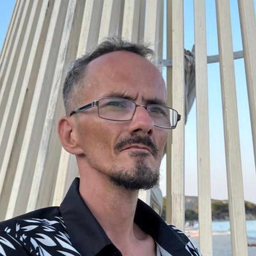 Ein Mann mit Brille und Ziegenbart steht vor hölzernen Latten am Strand.