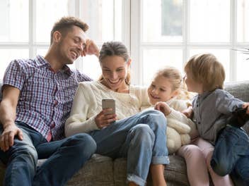 Junge Familie sitzt gemeinsam auf der Couch und schaut lächelnd auf ein Smartphone