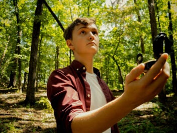 Ein Junge hält ein Kompass in der Hand, schaut darüber hinweg und steht in einem Wald
