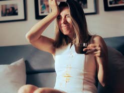 Eine Frau auf einem Sofa, die sich mit Kaffee besudelt hat und sich den Kopf hält.