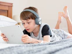 Ein Junge liegt auf einem Bett, trägt Kopfhörer und schaut auf ein Tablet