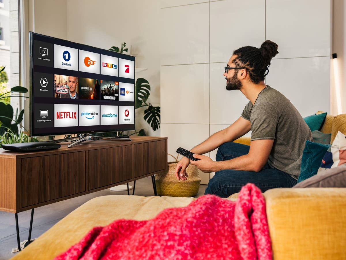 Ein Mann sitzt auf einer Couch im Wohnzimmer. Auf dem Fernseher ist Magenta TV zu sehen