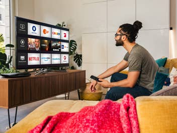 Ein Mann sitzt auf einer Couch im Wohnzimmer. Auf dem Fernseher ist Magenta TV zu sehen