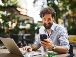 Mann sitzt an einem Laptop und schaut in sein Handy