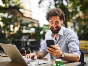 Mann sitzt an einem Laptop und schaut in sein Handy