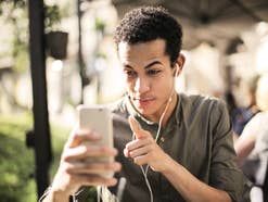 Junger Mann hat einen Videoanruf auf seinem Handy Ein junger Mann mit schwarzen Locken redet auf sein Smartphone ein und hat Kopfhörer in den Ohren