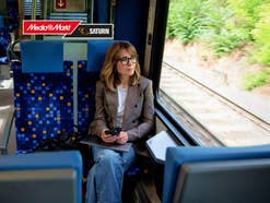 Eine Frau sitzt in einem Zug und schaut aus dem Fenster. In der Ecke oben links ist ein MediaMarkt-Logo.
