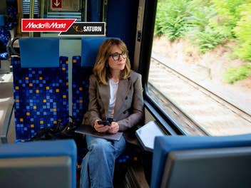 Eine Frau sitzt in einem Zug und schaut aus dem Fenster. In der Ecke oben links ist ein MediaMarkt-Logo.
