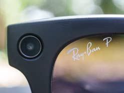 Nahaufnahme der oberen Ecke einer schwarzen Ray-Ban-Smart-Glasses, auf der das kreisrunde Kamera-Objektiv im Rahmen sowie der Schriftzug 