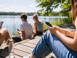 Jugendliche sitzen auf einem Steg am See und nutzen ihre Smartphones
