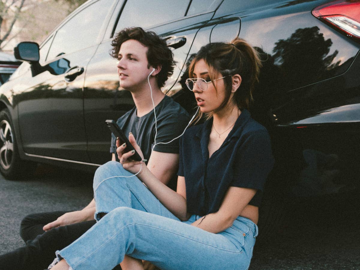 Musik-Streaming-Dienste im Vergleich im V Ein Mann und eine Frau sitzen vor einem Auto und hören über Kopfhörer Musik.