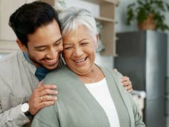 Ein Mann hält eine ältere Frau im Arm. Beide stehen in einer Wohnung und lachen