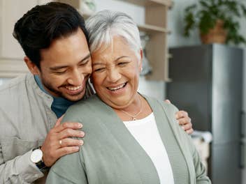 Ein Mann hält eine ältere Frau im Arm. Beide stehen in einer Wohnung und lachen