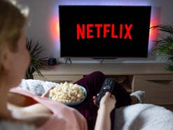 Frau sitz mit Popcorn und Fernbedienung auf der Couch und schaltet Netflix auf ihrem TV ein.