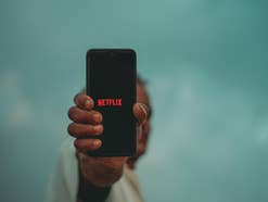 Das Netflix-Logo auf einem Handy, das eine Person in der Hand und in die Kamera hält.