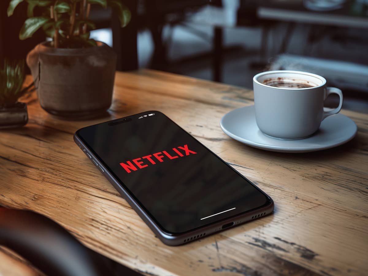 Das Netflix-Logo ist auf einem Display eines Smartphones abgebildert, das auf einem Holztisch neben einer Tasse Kaffee liegt.