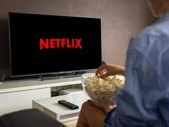 Mannschaut mit Popcorn in der Hand auf einen Fernseher mit Netflix-Logo.