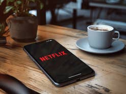Das Netflix-Logo ist auf einem Display eines Smartphones abgebildert, das auf einem Holztisch neben einer Tasse Kaffee liegt.