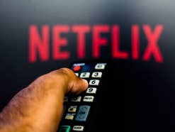 Netflix Logo im Hintergrund, während Mann eine Fernbedienung in der Hand hält.