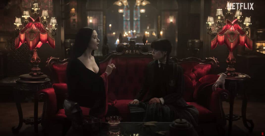 Morticia Addams sitzt auf einer roten Couch und spricht mit Wednesday in einer gotischen Umgebung. Netflix-Logo sichtbar.