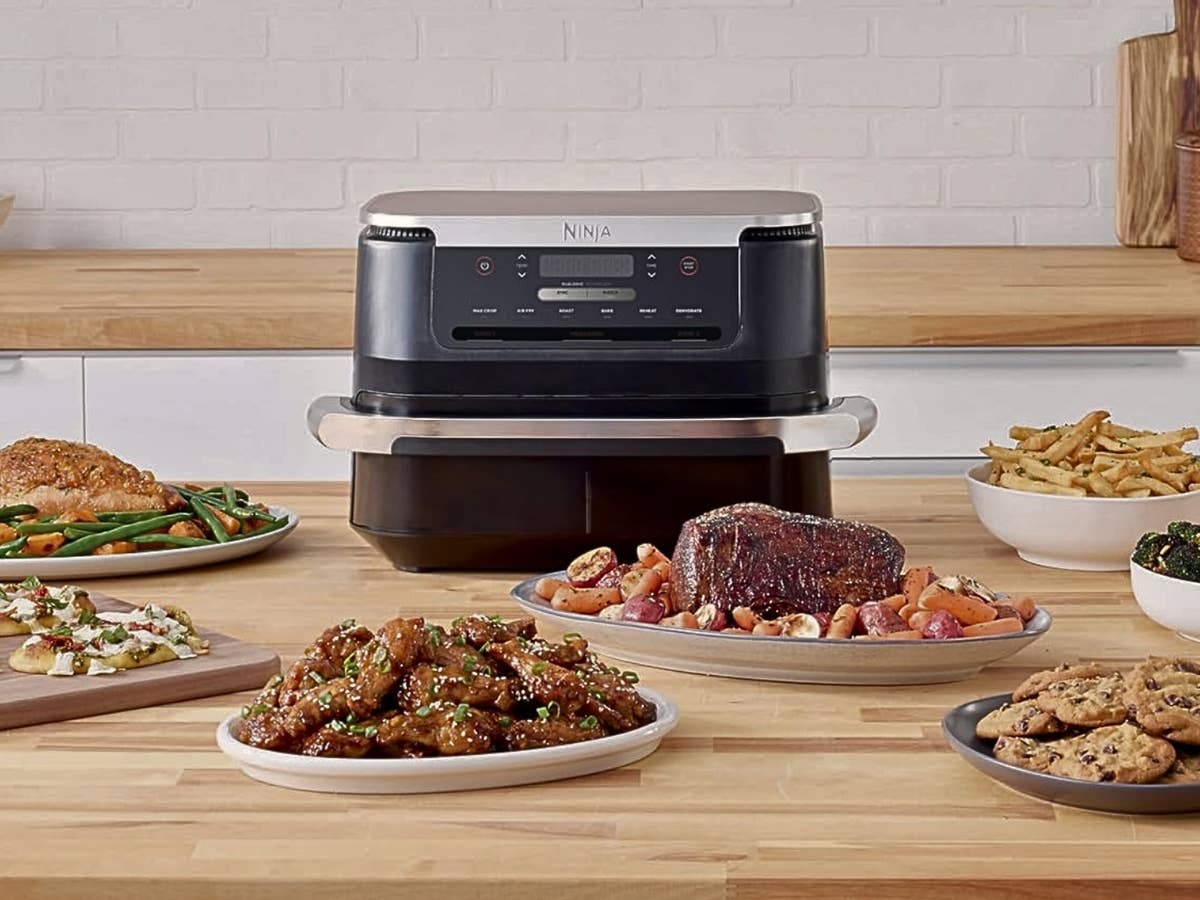 Ninja FlexDrawer Airfryer bei Amazon im Angebot Ein Airfryer von Ninja steht in der Mitte zwischen verschiedenen Tellern mit Essen auf einem Tisch.