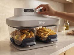 Ninja Crispi DualZone Heißluftfritteuse gefüllt mit Pommes und Hähnchen, eine Hand bedient das Gerät und dreht den großen Regler zwischen den beiden Displays