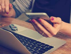 EIn Laptop steht auf einem Tisch, eine Frau hält ein iPhone in der Hand
