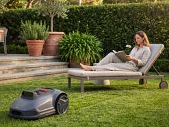 Eine Frau sitzt auf einer Liege im Garten, ein Rasenmähroboter mäht