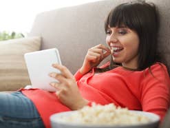 Frau streamt Videos auf einem Tablet und isst dabei Popcorn.