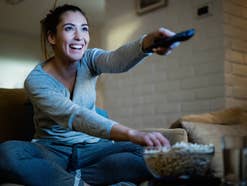 Frau sitzt mit einer Fernbedienung und Popcorn vor einem Fernseher.