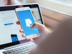 Eine Person hat einen Laptop geöffnet und ein Handy in der Hand. Auf beiden ist PayPal geöffnet.
