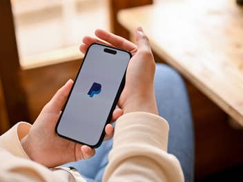 Frau sitzt vor einem Tisch am Fenster und hat ihr Smartphone in den Händen, auf dem die PayPal-App geöffnet ist