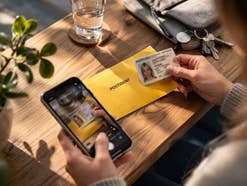 Eine Frau mit einem POSTIDENT-Umschlag, einem Smartphone und einem Personalausweis