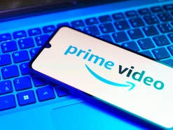 Logo von Prime Video auf einem Smartphone-Display das vor blauem Hintergrund auf einer Laptop-Tastatur liegt.