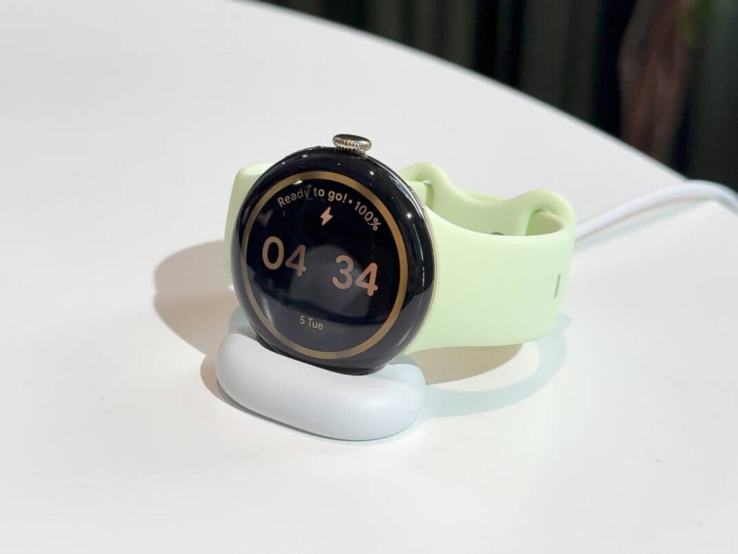 Eine Google Pixel Watch 4 auf einer Ladestation, die die Uhrzeit 04:34 und einen Akkustatus von 100% anzeigt.