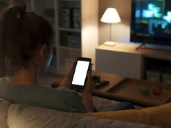 Ein Teenager sitzt auf einer Couch, hält ein Handy in der Hand. Im Hintergrund ist ein TV zu sehen.