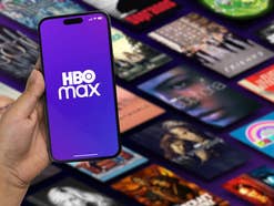 Jemand hält ein Smartphone in der Hand, auf dem das HBO Max-Logo zu sehen ist und im Hintergrund sieht man Einblendungen der Filme und Serien, die auf der Streamingplattform angeboten werden.